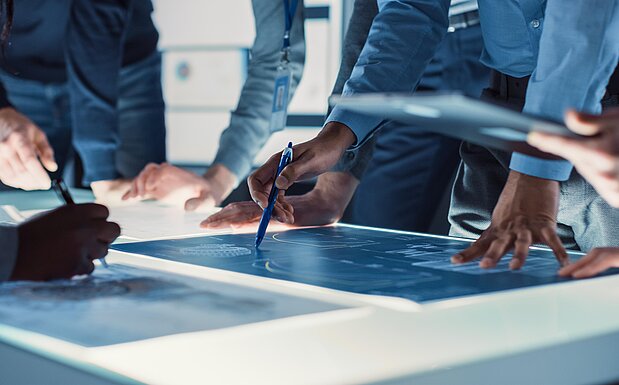 Mehrere Personen stehen um einen Tisch und arbeiten gemeinsam an Plänen oder technischen Zeichnungen