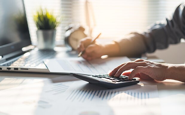 Person arbeitet am Schreibtisch mit Taschenrechner, Laptop und Notizen.